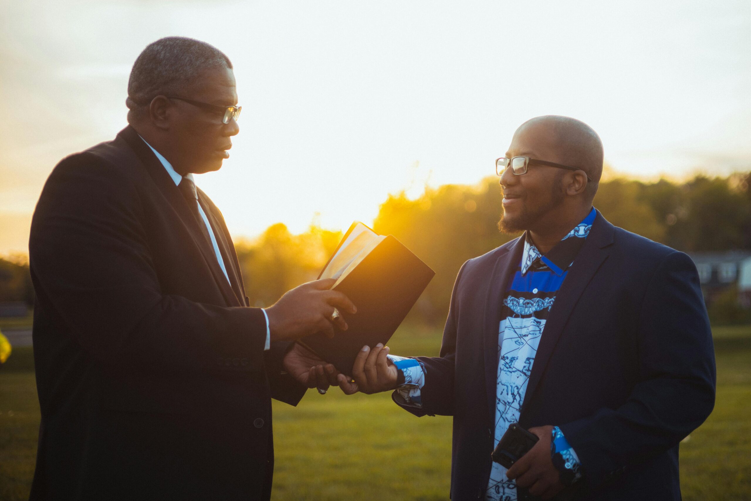 Two men sharing a moment with a book outdoors during sunset, symbolizing learning and togetherness.